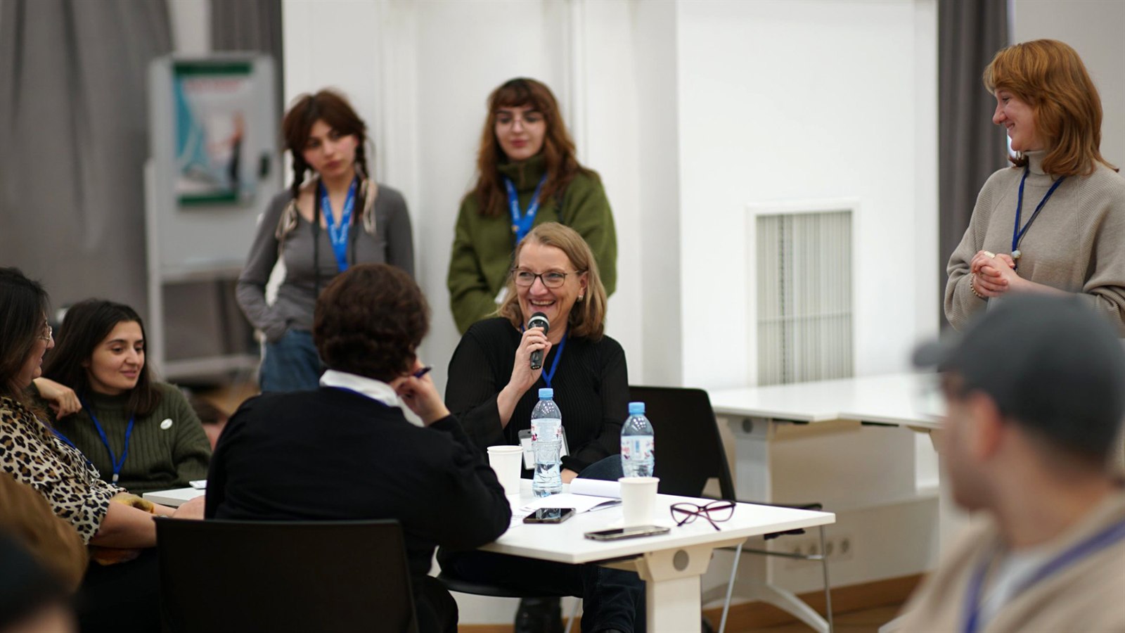 Barbara Weidmann spricht lächelnd mit einem Mikrofon in einer Workshoprunde; um sie herum sitzen und stehen Teilnehmende.